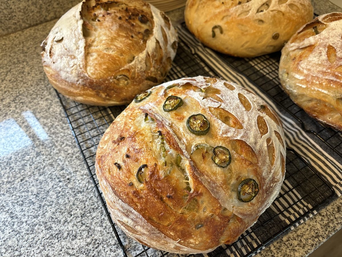 Multiple sourdough loaves with visible jalapenos cooling on wire rack, banneton baskets in background